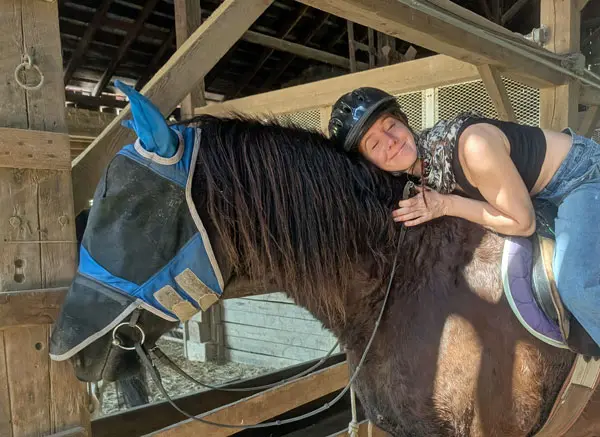woman wearing a helmet smiling and hugging a brown horse wearing a blue fly mask inside a barn stable.