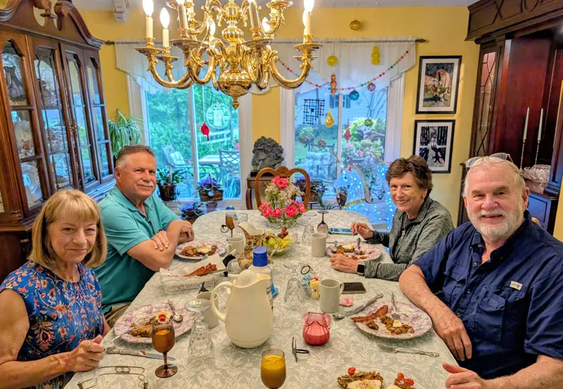 Four adults smiling while sitting around a dining table set for breakfast in a brightly lit room.